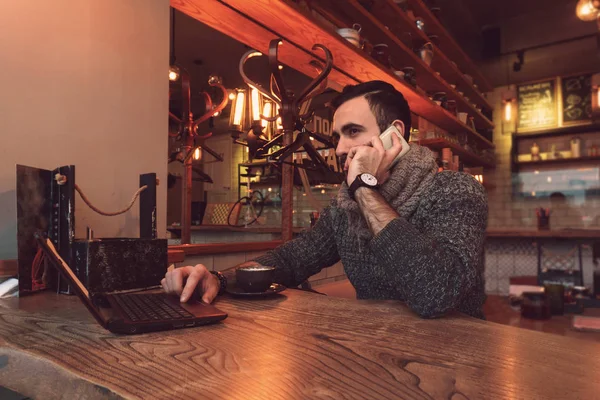 Un joven con barba se sienta en una mesa en un café y trabaja detrás de un pequeño portátil con una taza de delicioso café — Foto de Stock