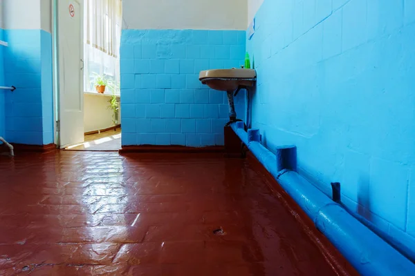 An old, untreated bathroom with one rusty sink and a leaking faucet and blue tiles on the wall