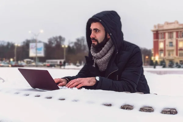 A young man with a beard in his hood works for a laptop in the winter in the snow when it\'s very cold outside