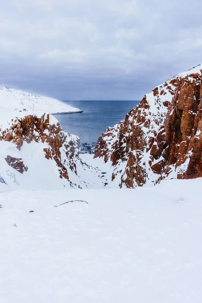Vážné a nebezpečné skály oranžové barvy na pobřeží oceánu na severu pod sněhem — Stock fotografie