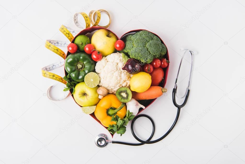 vegetables and fruits laying in heart shaped dish near stethoscope and measuring tape isolated on white background   
