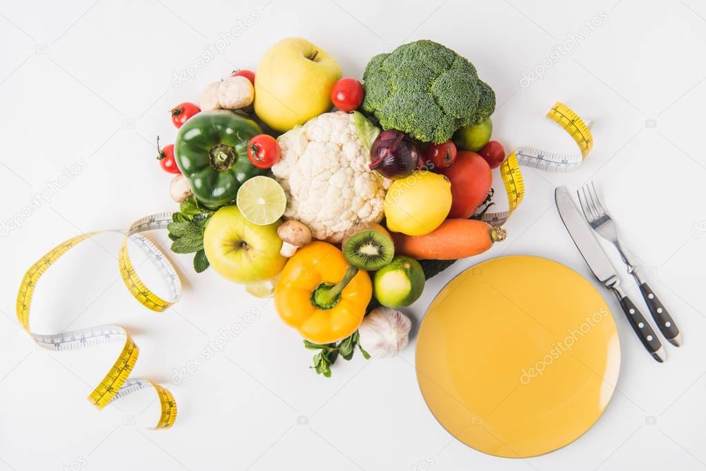 vegetables and fruits laying on white background with fork, spoon, measuring tape and plate