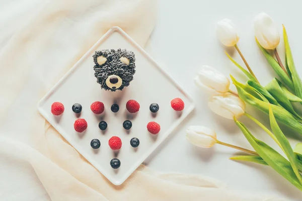 top view of sweet tasty muffin in shape of bear, fresh raspberries and tulip flowers  
