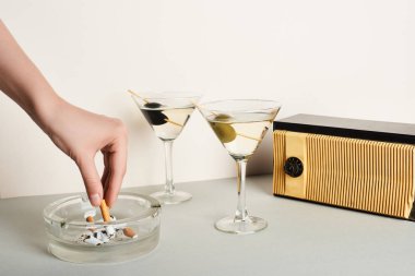 Cropped view of woman putting cigarette to ashtray beside cocktails and vintage radio on white background clipart
