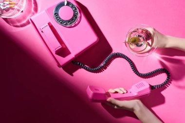 Pembe yüzey üzerinde sigara izmaritleri olan bir kadının elinde kokteyl ve telefon ahizesi.