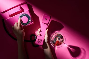 Pembe arka planda kokteylin yanında antika telefon kullanan bir kadın görüntüsü.