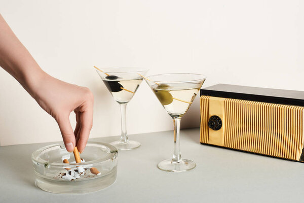 Cropped view of woman putting cigarette to ashtray beside cocktails and vintage radio on white background