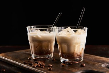 white russian cocktail in glasses with straws on wooden board with coffee grains isolated on black