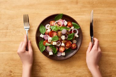 Ahşap masada çatal ve bıçakla panzanella salatası yiyen bir kadın görüntüsü.