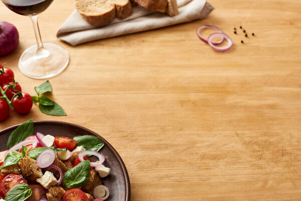 delicious Italian vegetable salad panzanella served on plate on wooden table near fresh ingredients, bread and red wine