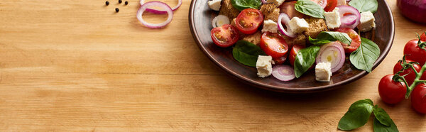 delicious Italian vegetable salad panzanella served on plate on wooden table near fresh ingredients, panoramic shot