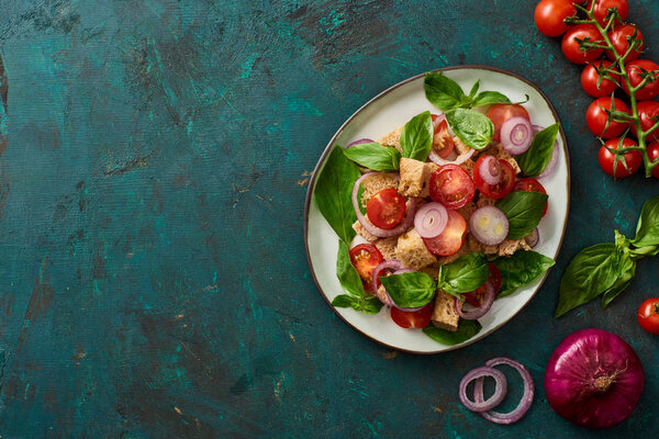 сверху вид на вкусный итальянский овощной салат panzanella подается на тарелке на текстурированной зеленой поверхности с помидорами, базиликом и красным луком
