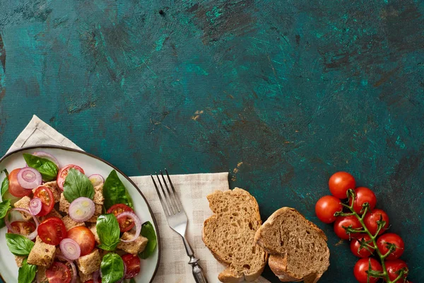 top view of delicious Italian vegetable salad panzanella served on plate on textured green surface with tomatoes, bread, napkin and fork