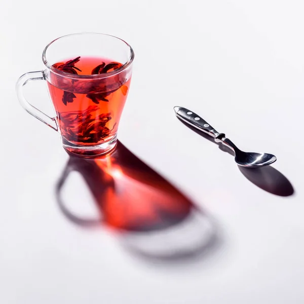 Taza de vidrio con té de hibisco y cuchara en la mesa blanca - foto de stock