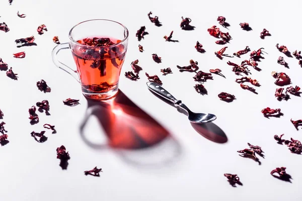 Tasse en verre de thé hibiscus avec cuillère et thé éparpillé sur la table — Photo de stock