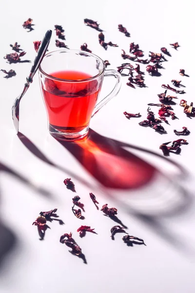 Tasse en verre de thé hibiscus avec cuillère et thé éparpillé sur la table — Photo de stock