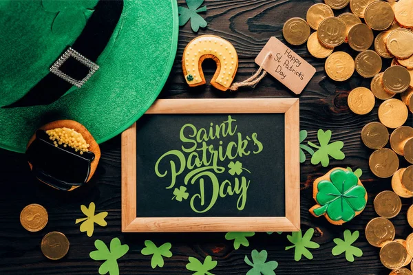 Flat lay with golden coins, festive cookies, hat and blackboard with saint patricks day lettering on wooden surface — Stock Photo