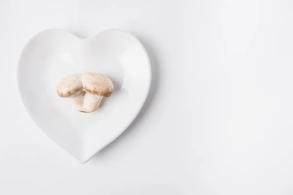 Cogumelos crus que colocam na placa em forma de coração no fundo branco — Fotografia de Stock