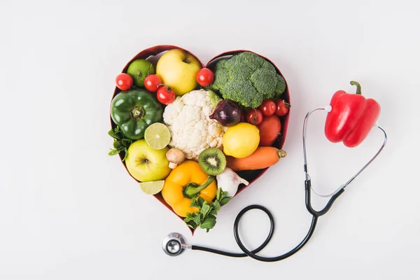 Vegetables and fruits laying in heart shaped dish near pepper with stethoscope isolated on white background — Stock Photo