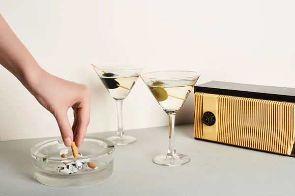 Обрезанный вид женщины, кладущей сигарету в пепельницу рядом с коктейлями и винтажным радио на белом фоне — стоковое фото