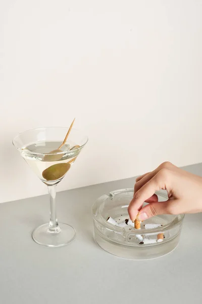Vista cortada da mulher colocando cigarro no cinzeiro com coquetel martini na superfície cinza — Fotografia de Stock