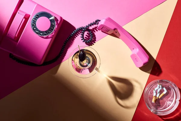 Vista dall'alto telefono vintage con martini in vetro e posacenere con mozziconi di sigaretta su superficie colorata — Foto stock