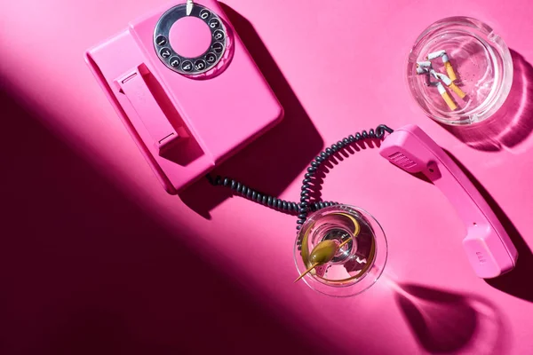 Vista dall'alto di cocktail e telefono retrò accanto a smarrito con mozziconi di sigaretta sulla superficie rosa — Foto stock