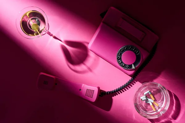 Top view of martini, pink telephone and astray with cigarette butts on pink surface — Stock Photo