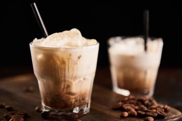 Selective focus of white russian cocktail in glasses with straws on wooden board with coffee grains isolated on black — Stock Photo