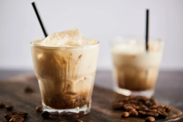 Selective focus of white russian cocktail in glasses with straws on wooden board with coffee grains isolated on grey — Stock Photo