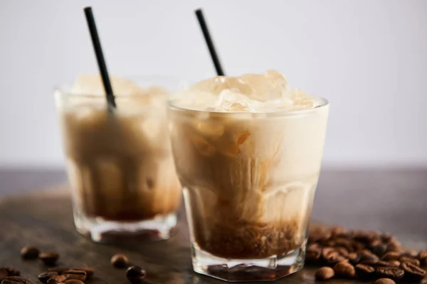 Selective focus of white russian cocktail in glasses with straws on wooden board with coffee grains isolated on grey — Stock Photo