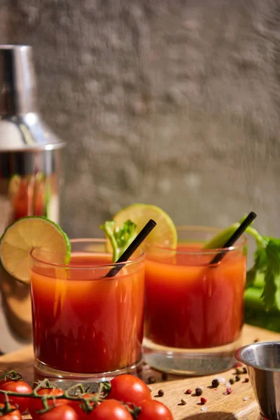 Кровавый коктейль в стаканах с соломинкой и лаймом на деревянной доске рядом с солью, перцем, помидорами и сельдереем — стоковое фото