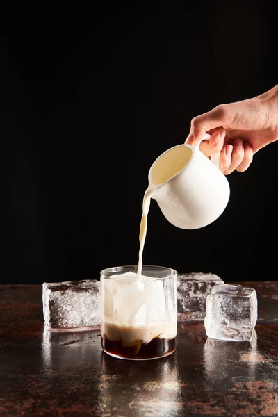 Vista recortada de la mujer poring leche en vidrio con cubitos de hielo aislados en negro - foto de stock