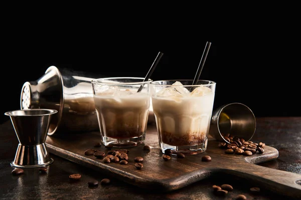 Белый русский коктейль в стаканах с соломинками на деревянной доске с зерном кофе и шейкером, изолированным на черном — стоковое фото
