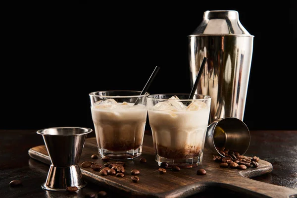 White russian cocktail in glasses with straws on wooden board with coffee grains and shaker isolated on black — Stock Photo