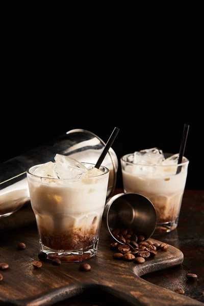 White russian cocktail in glasses with straws on wooden board with coffee grains and shaker isolated on black — Stock Photo