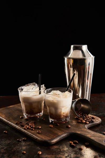 Белый русский коктейль в стаканах с соломинками на деревянной доске с зерном кофе и шейкером, изолированным на черном — стоковое фото
