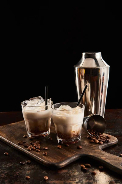 White russian cocktail in glasses with straws on wooden board with coffee grains and shaker isolated on black — Stock Photo