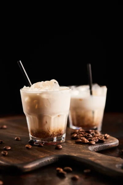 White russian cocktail in glasses with straws on wooden board with coffee grains isolated on black — Stock Photo