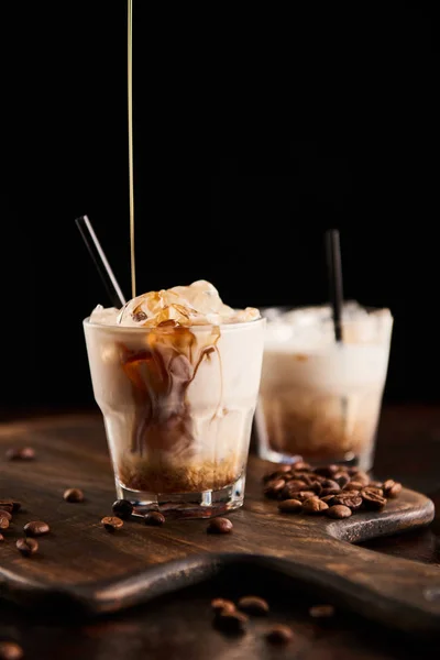 Licor de café vertiendo en blanco cóctel ruso en vasos con pajitas sobre tabla de madera con granos de café aislados en negro - foto de stock