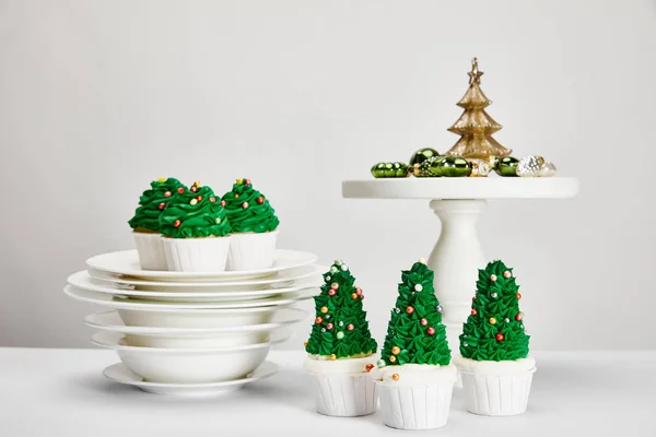 Deliciosos cupcakes de árbol de Navidad con platos y bolas brillantes en la superficie blanca aislada en gris - foto de stock
