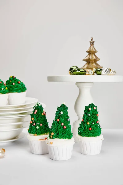 Leckere Weihnachtsbaum-Cupcakes mit Tellern und glänzenden Kugeln auf weißer Oberfläche isoliert auf grau — Stockfoto