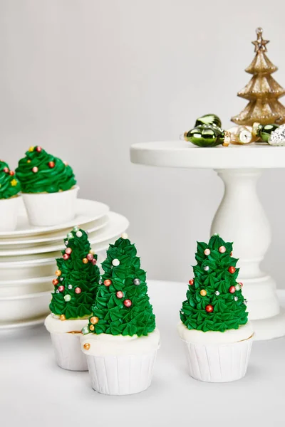 Deliciosos cupcakes de árbol de Navidad con platos y bolas brillantes en la superficie blanca aislada en gris - foto de stock