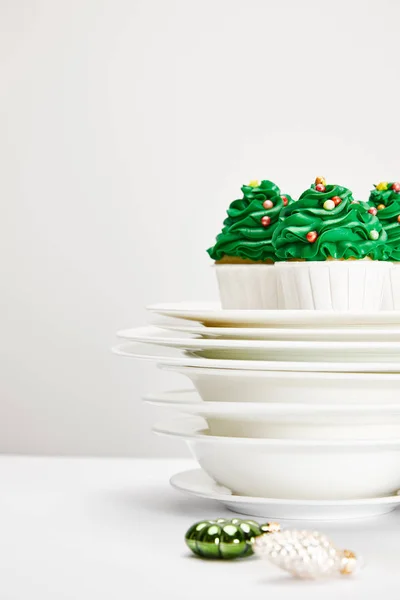 Selektiver Fokus von leckeren Weihnachtsbaum-Cupcakes mit Tellern und glänzenden Kugeln auf weißer Oberfläche isoliert auf grau — Stockfoto