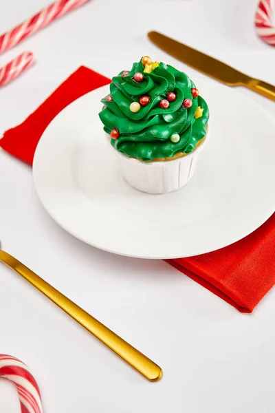 Delicioso cupcake en plato blanco con cubiertos de oro, caramelos y servilleta roja en la superficie blanca - foto de stock