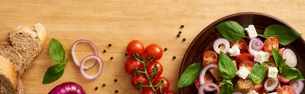 Vista superior de la deliciosa ensalada de verduras italiana panzanella servido en el plato en la mesa de madera cerca de ingredientes frescos, tiro panorámico - foto de stock