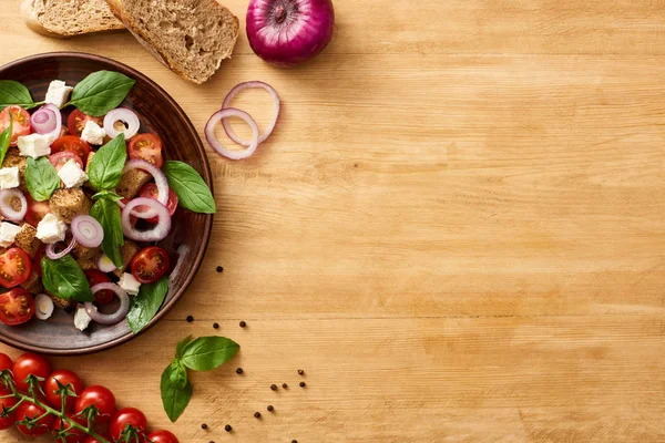 Сверху вид на вкусный итальянский овощной салат panzanella подается на тарелке на деревянный стол рядом со свежими ингредиентами — стоковое фото