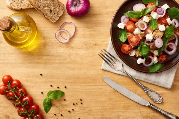 Vista superior de la deliciosa ensalada de verduras italiana panzanella servido en el plato en la mesa de madera cerca de ingredientes frescos y cubiertos — Stock Photo