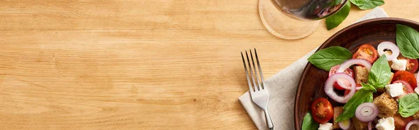 Top view of delicious Italian vegetable salad panzanella served on plate on wooden table near fork on napkin and red wine, panoramic shot — Stock Photo