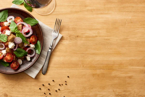 Vista superior de la deliciosa ensalada de verduras italiana panzanella servido en el plato en la mesa de madera cerca de tenedor en servilleta y vino tinto - foto de stock